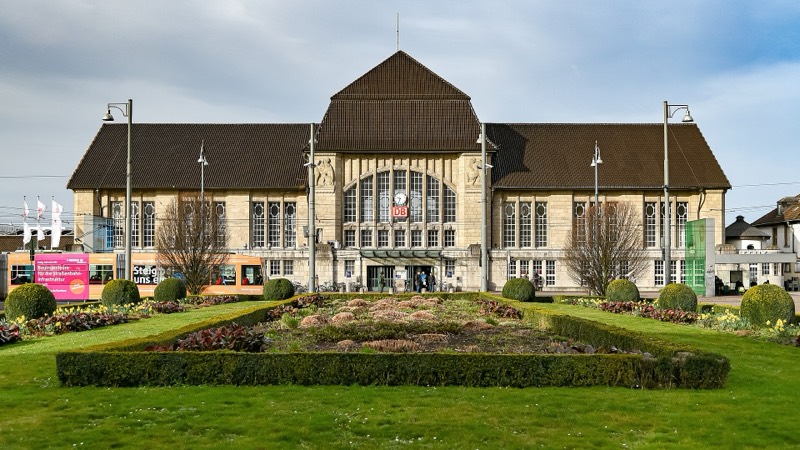 Empfangsgebäude des Darmstädter Hauptbahnhofs mit Grünanlage auf dem Vorplatz, links eine Straßenbahn