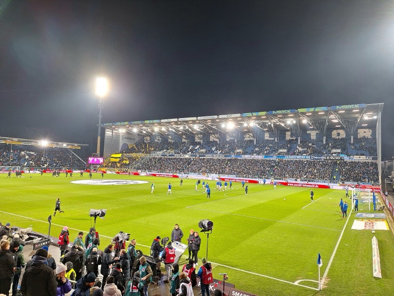 Flutlichtspiel im Stadion am Böllenfalltor: Blick über den Rasen auf die vollbesetzte Gegengerade beim Aufwärmen vor dem Spiel