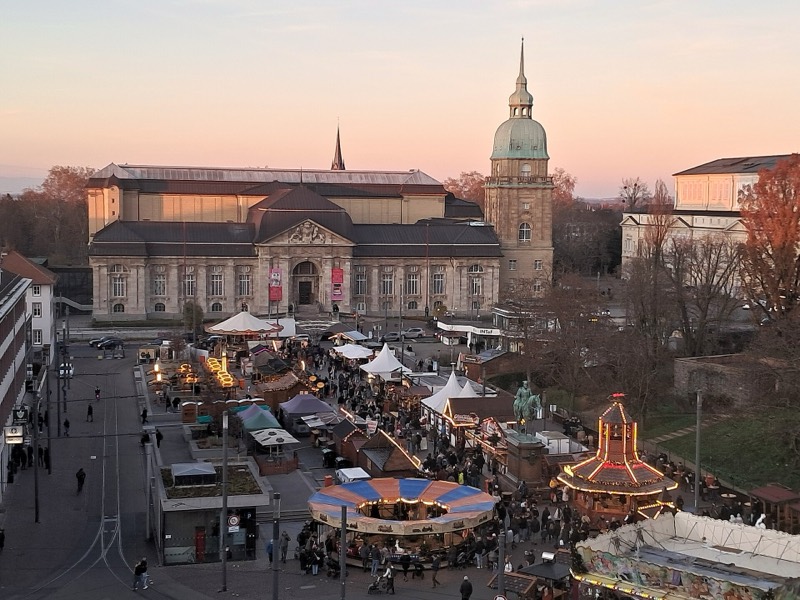 Weihnachtsmarkt auf dem Darmstädter Friedensplatz in der Abenddämmerung mit Karussell, beleuchteten Ständen und dem Hessischen Landesmuseum im Hintergrund